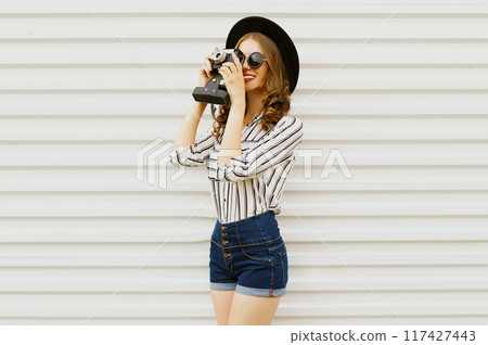 Happy smiling young woman holding camera in black hat, white striped shirt on white wall background 117427443