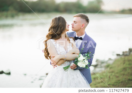Beautifull wedding couple kissing and embracing near the shore of a mountain river with stones Beautifull wedding couple kissing and embracing near the shore of a mountain river with stones 117427479