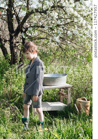 a little boy in shorts and a jacket waters plants from a watering can. Fashionable look for a country boy 117427957