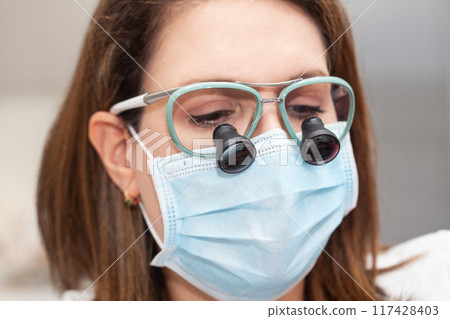 Close up of a female dermatologist face while suturing a patient during a small surgery wearing a binocular loupe. 117428403