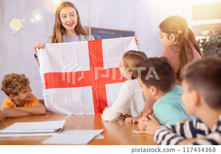 Young female teacher showing flag of England to schoolchildren preteens during history lesson in classroom Young female teacher showing flag of England to schoolchildren preteens during history lesson in classroom 117434368