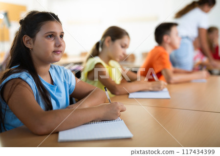 Interested preteen girl studying in classroom with classmates Interested preteen girl studying in classroom with classmates 117434399