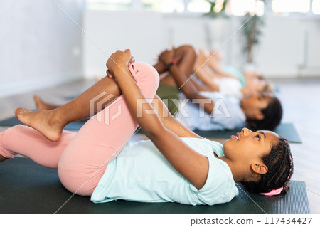 Children doing yoga with group in studio, lying on mat with knee pressed to chest with hands, performing hamstring and low back stretching Ardha Pawanmuktasana Children doing yoga with group in studio, lying on mat with knee pressed to chest with hands, performing hamstring and low back stretching Ardha Pawanmuktasana 117434427