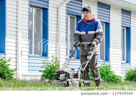 A man in a jacket and hat is using a lawn mower on the grass in front of a residential blue and 117434544