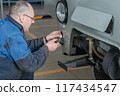 A mechanic in blue workwear installs a tow hitch on a white car in an automotive workshop 117434547