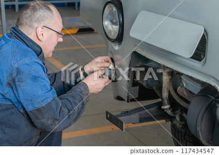 A mechanic in blue workwear installs a tow hitch on a white car in an automotive workshop 117434547