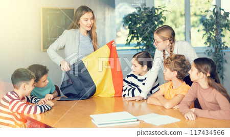 Smiling young female teacher explaining culture of country while showing flag of Belgium to schoolchildren preteens in classroom Smiling young female teacher explaining culture of country while showing flag of Belgium to schoolchildren preteens in classroom 117434566
