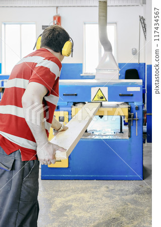 A worker in a workshop uses a large industrial woodworking machine to process a wooden plank 117434567