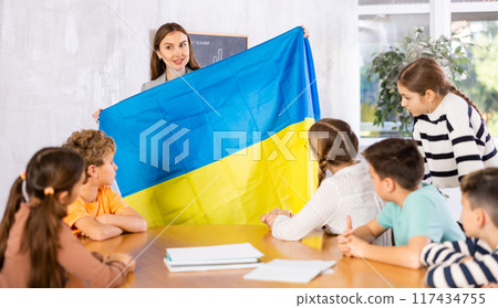 Smiling young woman teacher showing national flag of Ukraine and telling preteens schoolchildren history of country during lesson in class Smiling young woman teacher showing national flag of Ukraine and telling preteens schoolchildren history of country during lesson in class 117434755