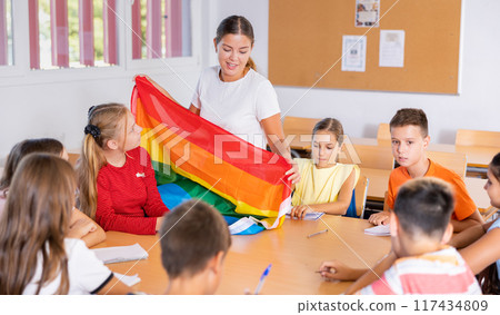 Children sitting in classroom and listening to female teacher. She holding rainbow flag in hands and talking about minorities 117434809
