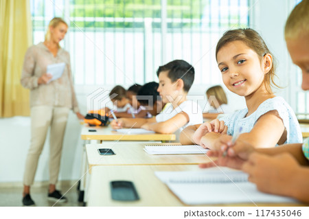 Girl sits at a desk next to diligent elementary school students Girl sits at a desk next to diligent elementary school students 117435046