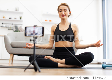 Woman practicing yoga at home while recording video for online class Woman practicing yoga at home while recording video for online class 117437000
