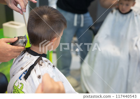 Barber trimming boy's hair from back wearing protective cape in salon Barber trimming boy's hair from back wearing protective cape in salon 117440543