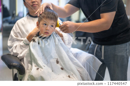 Child getting a haircut while sitting on a caretaker's lap in a salon 117440547