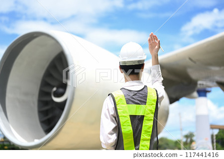 Airport worker in a safety vest standing in front of a large airplane on runway Airport worker in a safety vest standing in front of a large airplane on runway 117441416