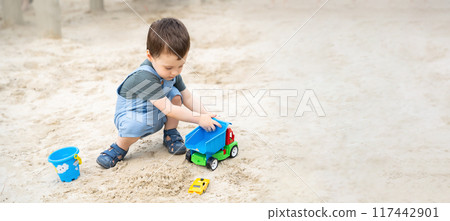 Little cute toddler boy 2.5 years old plays in the sandbox on a sunny summer day. Outdoor creative activities for kids. Toy car and shovel. Selective focus. banner 117442901