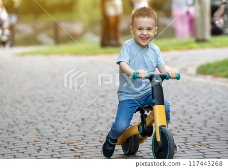 A cute happy caucasian toddler boy of three or four years old rides a bicycle or balance bike in the park against the backdrop of a crowd of people in the background. Summer activity for children. A cute happy caucasian toddler boy of three or four years old rides a bicycle or balance bike in the park against the backdrop of a crowd of people in the background. Summer activity for children. 117443268