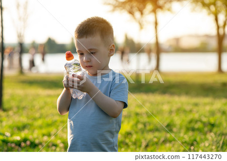 A little caucasian toddler boy of three and a half years old is standing in a summer public park with a bottle of clean water. Childhood and nature. Portrait A little caucasian toddler boy of three and a half years old is standing in a summer public park with a bottle of clean water. Childhood and nature. Portrait 117443270