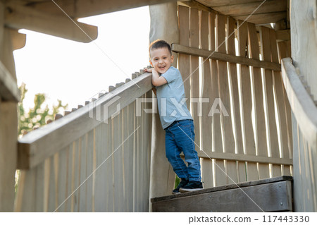 A cute happy three and half year old caucasian boy stands on a wooden ladder on the playground A cute happy three and half year old caucasian boy stands on a wooden ladder on the playground 117443330