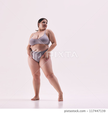 Full length photo of young overweight lady, model plus size posing in grey underwear highlights her shapes against white studio background. 117447139
