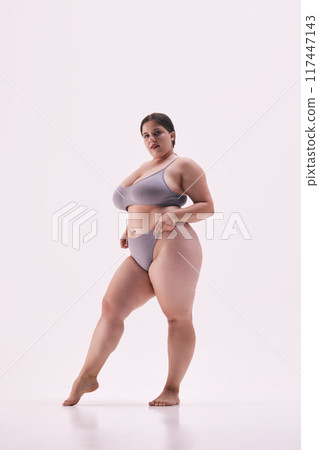 Chubby young lady posing shirtless showing her confidence against white studio background. Model dressed grey lingerie. Chubby young lady posing shirtless showing her confidence against white studio background. Model dressed grey lingerie. 117447143