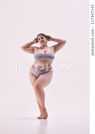 Full length photo of young overweight lady, model plus size posing in grey lingerie highlights her shapes against white studio background. 117447145