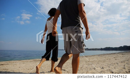 Couple holding hands of each other and walking at coast. Happy pair going along seashore at sunny day. Lovers spending time together at resort. Concept of vacation or holiday. Slow mo Couple holding hands of each other and walking at coast. Happy pair going along seashore at sunny day. Lovers spending time together at resort. Concept of vacation or holiday. Slow mo 117447412