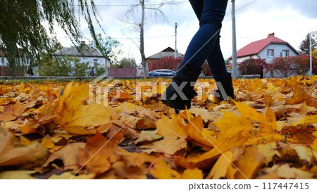 Legs of young woman stepping on yellow foliage at park. Female feet in boots going on fallen maple leaves at parkland. Girl walking at parkland at autumn season on cloudy day. Close up Slow motion 117447415