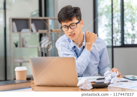 Tired and overworked young businessman using his hand to massage his sore neck while working late at the office Tired and overworked young businessman using his hand to massage his sore neck while working late at the office 117447837