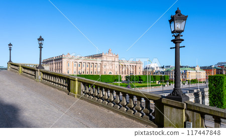 The Swedish House of Parliament in Stockholm basks in sunshine, with clear skies above and street lamps lining the adjacent bridge. Stockholm, Sweden The Swedish House of Parliament in Stockholm basks in sunshine, with clear skies above and street lamps lining the adjacent bridge. Stockholm, Sweden 117447848