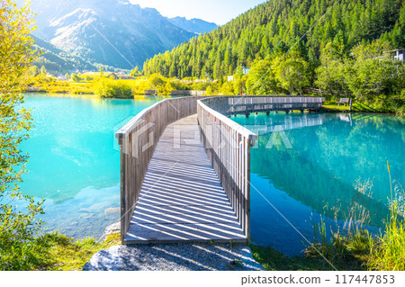 A tranquil wooden boardwalk bridge curves above the vibrant blue waters of Lake in Sulden, Italy. With the majestic Italian Alps towering in the background under a clear azure sky. 117447853