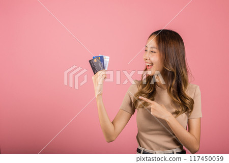 Portrait of Asian young woman holding bank card solution in hands in studio shot isolated on pink background, female smiling pointing and showing presenting credit card for online payment 117450059