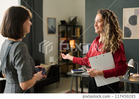 Two women engaging in a conversation inside modern office space with one woman holding a document and gesturing toward the other 117450285