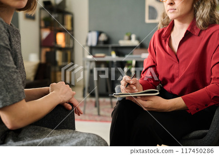 Woman wearing red shirt holding pen while talking with patient during session. Background consists of office furniture and bookshelf creating a warm and professional atmosphere 117450286