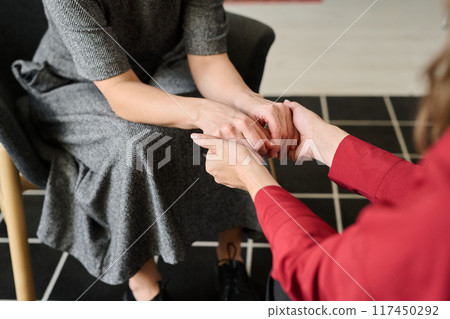 Comforting scene showing supportive gesture by two people sitting indoors with one person gently holding hands of the other, offering reassurance and warmth 117450292