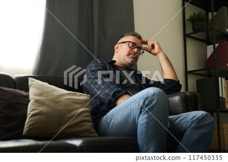 Man relaxing on couch in living room, resting head on hand while considering thoughts with serious expression near window with natural light coming through curtain Man relaxing on couch in living room, resting head on hand while considering thoughts with serious expression near window with natural light coming through curtain 117450335
