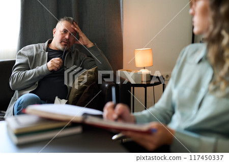 Man sitting on sofa contemplating while talking with therapist during therapy session indoors cozy room featuring comfortable sofa and lamp on side table 117450337