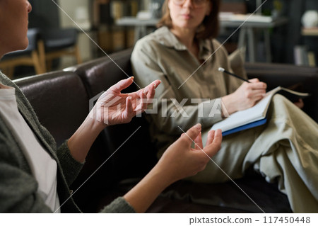 Two individuals engaging in counseling session on couch, one holding clipboard and pen, focusing on discussion, expressing thoughts with hand gestures Two individuals engaging in counseling session on couch, one holding clipboard and pen, focusing on discussion, expressing thoughts with hand gestures 117450448