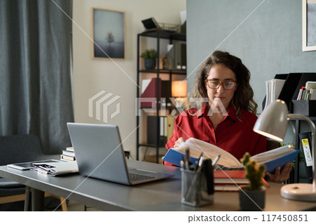 Focused woman with curly hair working at office desk with laptop and documents, surrounded by organized shelves and soft lighting, creating a productive workspace without interruptions 117450851