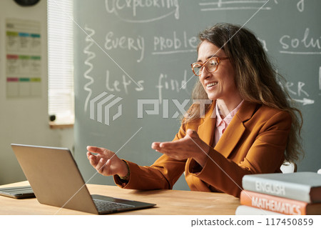Side view portrait of smiling female teacher talking to video chat during online lesson with student and gesturing 117450859