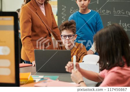 Colorful portrait of funny young boy wearing glasses and using laptop with group of children in school classroom copy space 117450867