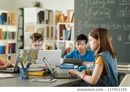 Side view portrait of young girl with prosthetic arm using laptop with inclusive group of children in school library copy space 117451105