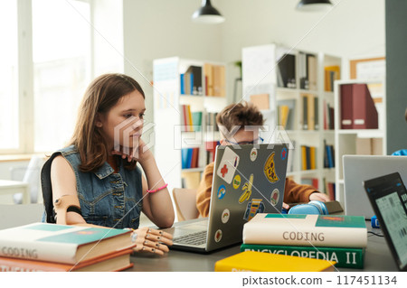 Portrait of young girl with arm prosthetic using laptop in modern school library while studying IT and programming copy space 117451134