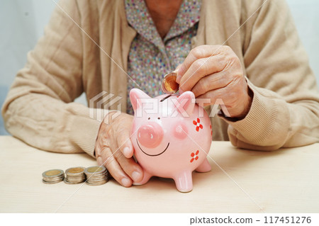Retired elderly woman putting coins money in piggy bank and worry about monthly expenses and treatment fee payment. 117451276