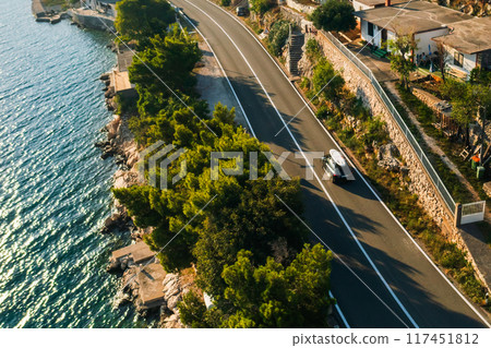 A sporty black car with a SUP on the roof races along a coastal road, the sun shining brightly overhead.  117451812