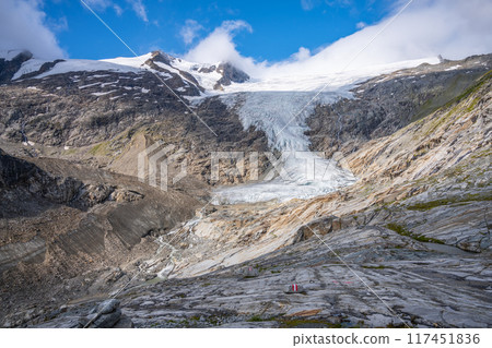 The Schlaten Glacier cascades down a rugged landscape, surrounded by rocky outcrops and patches of greenery. Bright blue sky and soft clouds frame the glacial expanse. 117451836