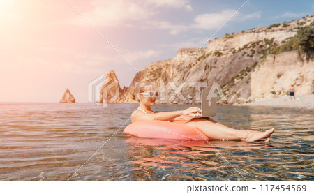 Woman freelancer works on laptop swimming in sea on pink inflatable ring. Happy tourist in sunglasses floating on inflatable donut and working on laptop computer in calm ocean. Remote working anywhere Woman freelancer works on laptop swimming in sea on pink inflatable ring. Happy tourist in sunglasses floating on inflatable donut and working on laptop computer in calm ocean. Remote working anywhere 117454569