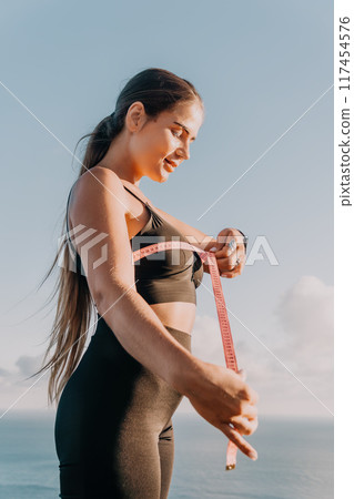 A woman is holding a pink tape measure and is measuring her breasts after morning fitness A woman is holding a pink tape measure and is measuring her breasts after morning fitness 117454576