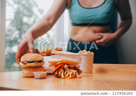 To eat or not. Cheerless plump woman looking at the burger while thinking to eat it or not To eat or not. Cheerless plump woman looking at the burger while thinking to eat it or not 117456163
