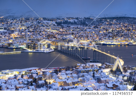 Panorama of Norwegian city of Troms in the winter. Snowy roofs, bridge, embankment near the port and fishing ships. Evening lights Panorama of Norwegian city of Troms in the winter. Snowy roofs, bridge, embankment near the port and fishing ships. Evening lights 117456557